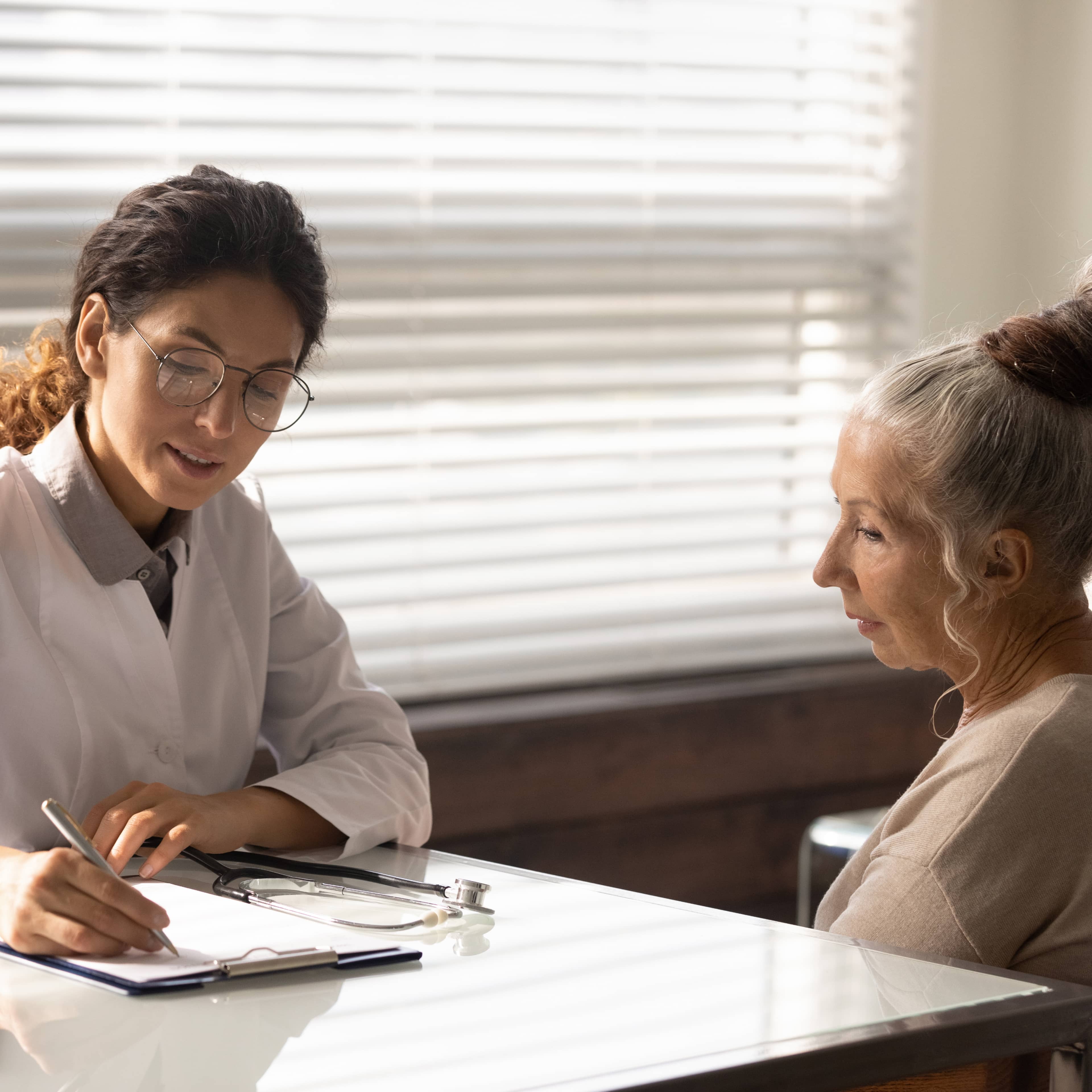 Médecin au travail avec des lunettes rédigeant un diagnostic des symptômes d'une patiente d'âge mûr dans un dossier médical.